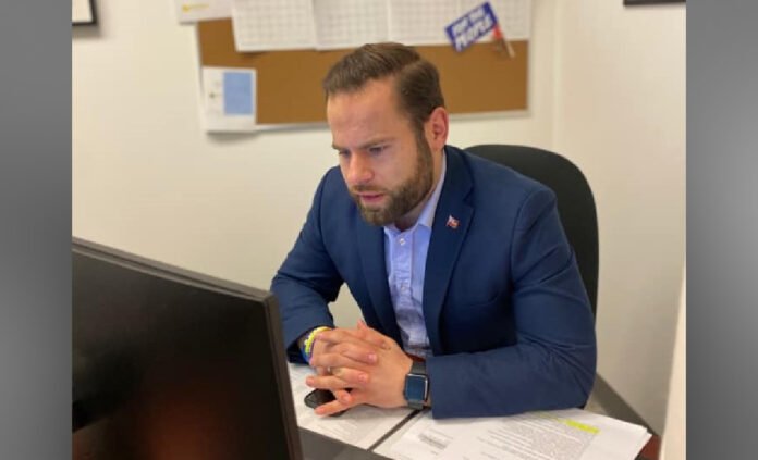 Ontario Labour Minister David Piccini sits at a table in a meeting room, wearing a blue suit and light blue shirt, hands clasped, with documents and a microphone in front of him.