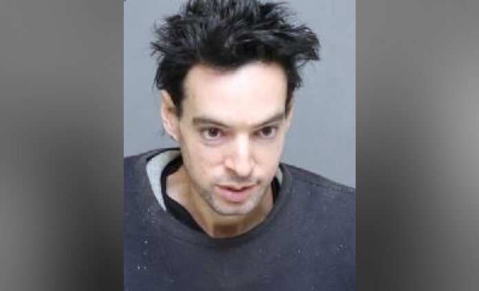 A head-and-shoulders police-released photograph of a man with short, dark, spiked hair, wearing a dark sweatshirt, photographed against a plain blue background.