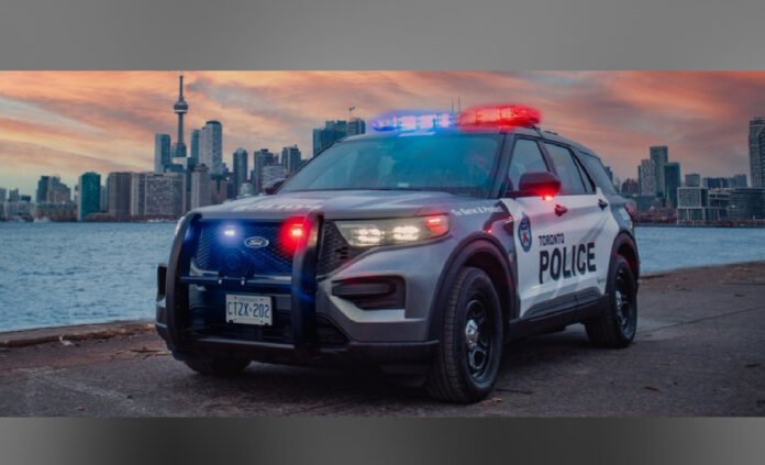 A Toronto Police Service cruiser with emergency lights flashing is parked along the waterfront at dusk, with the Toronto skyline and CN Tower visible in the background