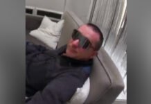 Toronto Police hunt suspect after multiple fires set at University of Toronto’s St. George Campus suspect ,a man wearing dark sunglasses reclines on a light-coloured couch or bed indoors, dressed in a dark jacket, with pillows and curtains visible in the background.