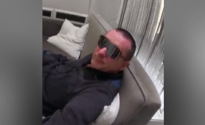 suspect ,a man wearing dark sunglasses reclines on a light-coloured couch or bed indoors, dressed in a dark jacket, with pillows and curtains visible in the background.