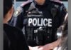 Vaughan commercial plaza shooting; second suspect now in police custody Close-up view of a York Regional Police officer wearing a black tactical vest with the word “POLICE” in large white letters across the front. The vest includes a radio, body camera, and shoulder patches, with the officer standing among other people.