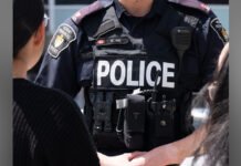 Vaughan commercial plaza shooting; second suspect now in police custody Close-up view of a York Regional Police officer wearing a black tactical vest with the word “POLICE” in large white letters across the front. The vest includes a radio, body camera, and shoulder patches, with the officer standing among other people.
