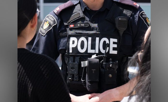 Close-up view of a York Regional Police  officer wearing a black tactical vest with the word “POLICE” in large white letters across the front. The vest includes a radio, body camera, and shoulder patches, with the officer standing among other people.