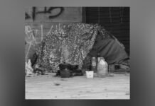 New report finds 85,000 Ontarians experienced homelessness last year Black-and-white image of a small homeless encampment set up against a wall. A blanket or tarp is draped over a makeshift shelter, with plastic bottles, cups, and containers placed on the ground in front. The scene suggests temporary living conditions in an urban setting.
