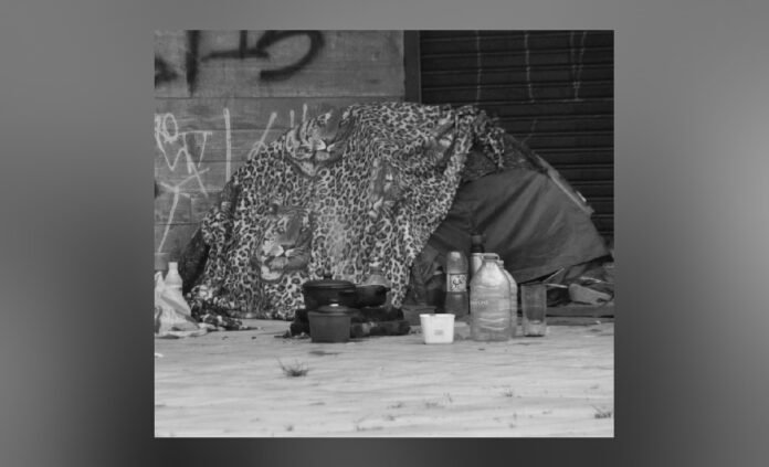 Black-and-white image of a small homeless encampment set up against a wall. A blanket or tarp is draped over a makeshift shelter, with plastic bottles, cups, and containers placed on the ground in front. The scene suggests temporary living conditions in an urban setting.