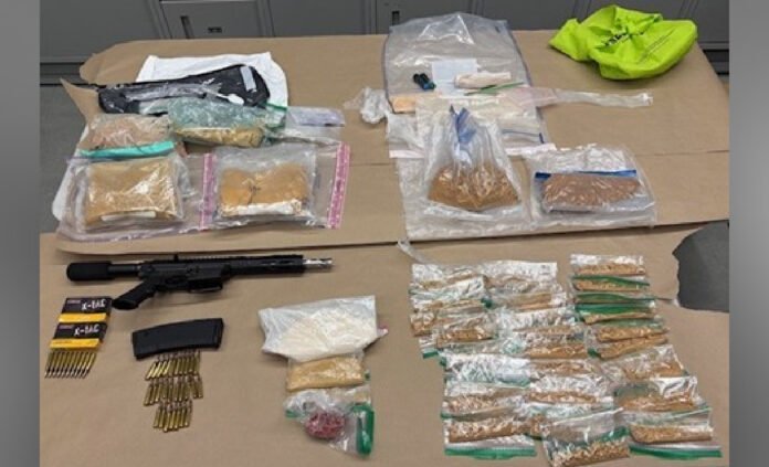 Photo shows police-seized evidence laid out on a table, including a rifle with magazine, boxes of ammunition, and numerous plastic bags containing suspected fentanyl for documentation.