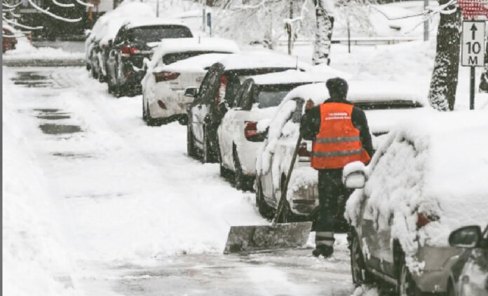 A person wearing an orange high-visibility vest shovels snow beside a row of cars parked along a residential street, with heavy snow covering the road, sidewalks, and vehicles after a winter storm.