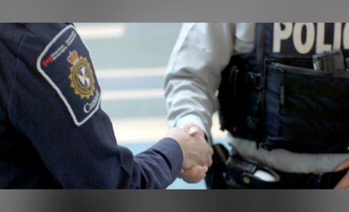 Sideways image of a uniformed officer with a visible Canada Border Services Agency shoulder patch shaking hand with a person wearing a grey hoodie, next to a dark police vehicle.