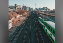 Metrolinx CEO apologizes to GO train riders for delays since train derailment A green-and-white commuter train travels along elevated railway tracks, with the tracks converging into the distance. In the background, Toronto’s skyline is visible, including the CN Tower, under an overcast sky.