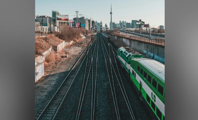 A green-and-white commuter train travels along elevated railway tracks, with the tracks converging into the distance. In the background, Toronto’s skyline is visible, including the CN Tower, under an overcast sky.