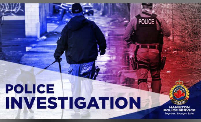 Hamilton Police A Hamilton Police Service graphic showing two police officers walking down a wet alleyway at night, illuminated by red and blue emergency lights. One officer wears a vest labeled “POLICE.” The words “POLICE INVESTIGATION” appear prominently, along with the Hamilton Police Service logo and the slogan