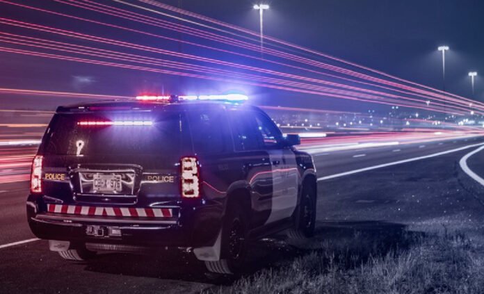 ntario Provincial Police cruiser with flashing red and blue lights parked on the shoulder of a highway at night, as long-exposure light trails from passing traffic streak past the scene.
