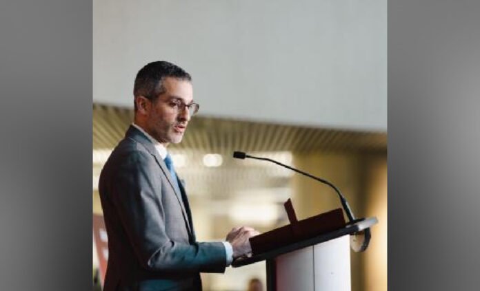 Inspector general of Ontario Ryan Teschner in a dark suit and tie speaks at a podium with a microphone, shown in profile, addressing an audience in an indoor conference setting.