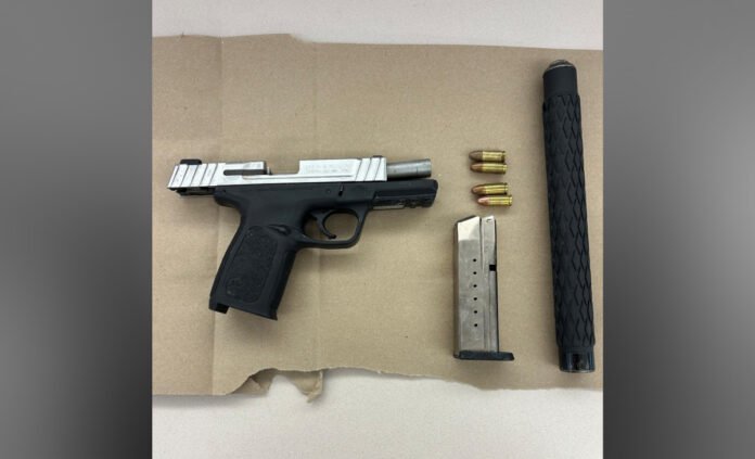 A black semi-automatic handgun with a silver slide is displayed on a cardboard surface. Below it lies a metal magazine, four loose bullets, and a long black cylindrical attachment believed to be a suppressor.