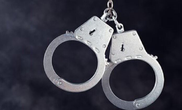 A close-up image of a pair of metal handcuffs with a short chain linking the two cuffs, shown against a dark background.