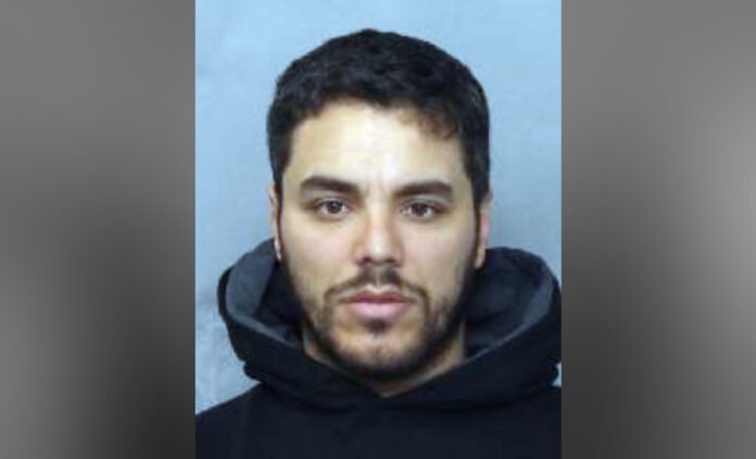 Portrait-style image of a man with short dark hair and a trimmed beard, wearing a dark hoodie, looking directly at the camera against a plain background.