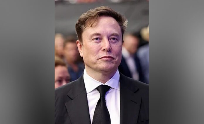 image of Elon Musk wearing a black suit, white shirt, and black tie stands facing the camera with a neutral expression. The background is blurred with several people visible behind him, suggesting a public or formal event setting.