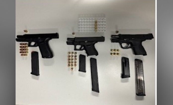 Three handguns displayed alongside multiple magazines and groups of ammunition, arranged on a white surface.