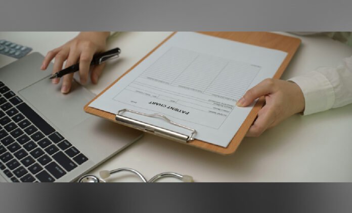 Hands holding a clipboard with a patient chart while another hand uses a pen next to a laptop and stethoscope on a desk.