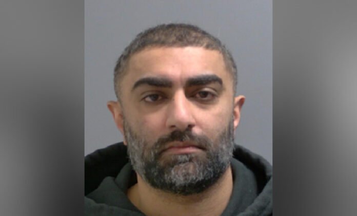 Headshot of man with short dark hair and a full beard with some grey. He has a medium complexion and is wearing a dark hoodie while looking straight at the camera against a plain light background.