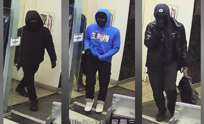 suspects_in_Markhamrobbery Three masked individuals captured on surveillance camera inside a building entrance, each wearing dark clothing; one in a blue hoodie and white shoes, another carrying a bag, and all appearing to conceal their identities.